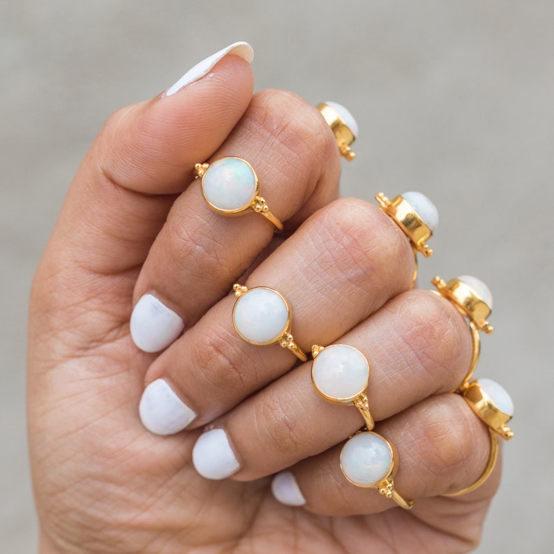 May include: A collection of gold-colored rings, each featuring a round, white opal stone. The rings are displayed on a hand with white nail polish. The rings have a simple, elegant design, with small decorative details on the band.