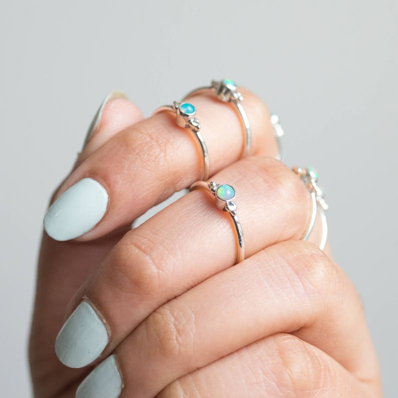 May include: A hand displays several silver rings, each featuring a small, round, opalescent gemstone. The rings have a delicate, minimalist design, with a thin band and subtle decorative elements near the gemstone. The nails are painted a light blue.