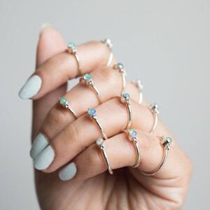 May include: A collection of silver rings, each featuring a small, round opal gemstone. The rings are displayed on a hand with light blue nail polish. The opals have a mix of colors, including blue, green, and white.