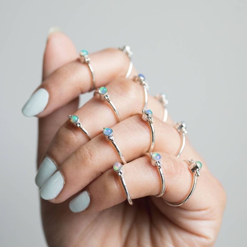May include: A collection of silver rings, each featuring a small, round opal gemstone. The rings are displayed on a hand with light blue nail polish. The opals have a mix of colors, including blue, green, and white.