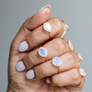 K&ouml;nnte beinhalten: Eine Hand zeigt mehrere goldene Ringe mit schillernden, oval geformten Mondsteinen. Die Ringe haben ein schlichtes, zartes Design, wobei die Steine in einem d&uuml;nnen Goldband gefasst sind. Der Hintergrund ist ein weiches, neutrales Grau.