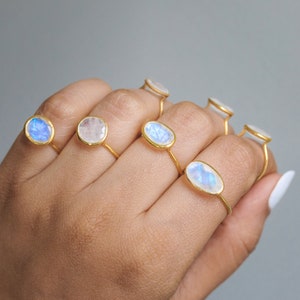 K&ouml;nnte beinhalten: Eine Sammlung goldener Ringe, jeder mit einem Mondstein in unterschiedlicher Form. Die Mondsteine zeigen eine Reihe von Farben, darunter Blau, Wei&szlig; und schillernde Farbt&ouml;ne. Die Ringe werden auf einer Hand vor einem neutralen grauen Hintergrund pr&auml;sentiert.