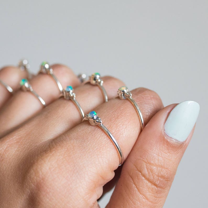 May include: A collection of silver rings, each featuring a small, iridescent opal gemstone. The rings are displayed on a hand with light skin, showcasing their delicate design. The opals display a range of colors, including blue, green, and white.