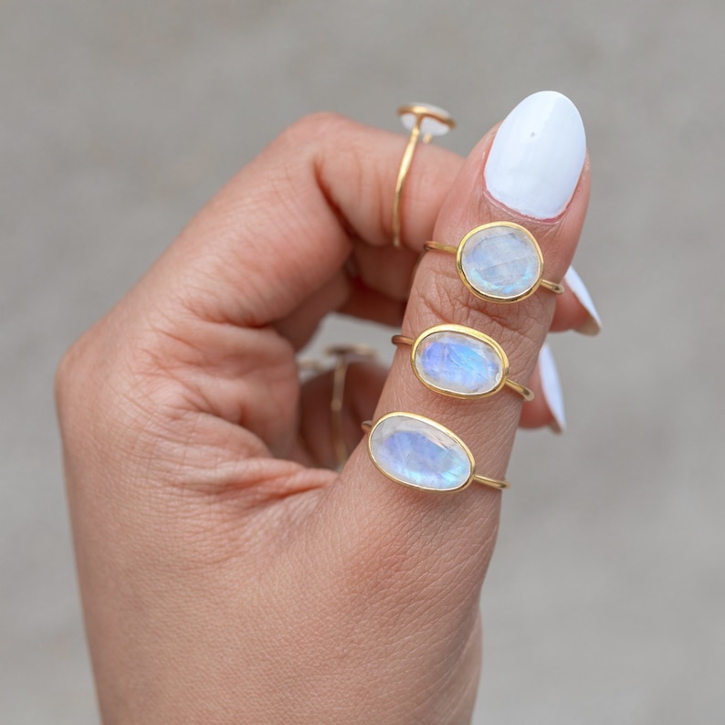 K&ouml;nnte beinhalten: Mehrere goldene Ringe mit ovalen, schillernden Mondsteinen werden auf einer Hand pr&auml;sentiert. Die Ringe haben ein zartes, d&uuml;nnes Band. Die Mondsteine haben eine blau-wei&szlig;e Farbe. Der Hintergrund ist ein neutrales Grau.