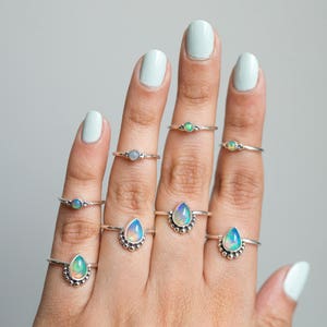 May include: A hand displays several silver rings with opalescent gemstones. The rings feature various designs, including round and teardrop shapes, and are set on delicate silver bands. The opals display a range of colors, including blue, green, and white.