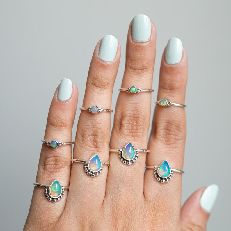 May include: A hand displays several silver rings with opalescent gemstones. The rings feature various designs, including round and teardrop shapes, and are set on delicate silver bands. The opals display a range of colors, including blue, green, and white.