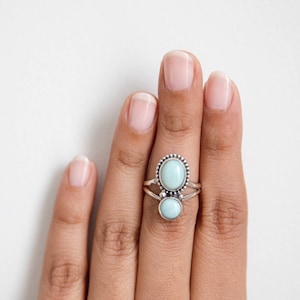 May include: A silver ring featuring two oval, light blue gemstones. The larger stone is set above a smaller one, with a beaded border around the top stone. The ring is displayed on a hand.
