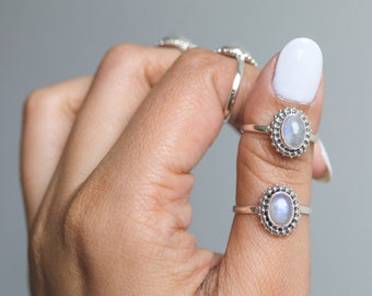 May include: Two silver rings with oval, iridescent moonstone cabochons are displayed on a hand. The rings have a beaded border around the moonstone. The background is a solid, light gray.