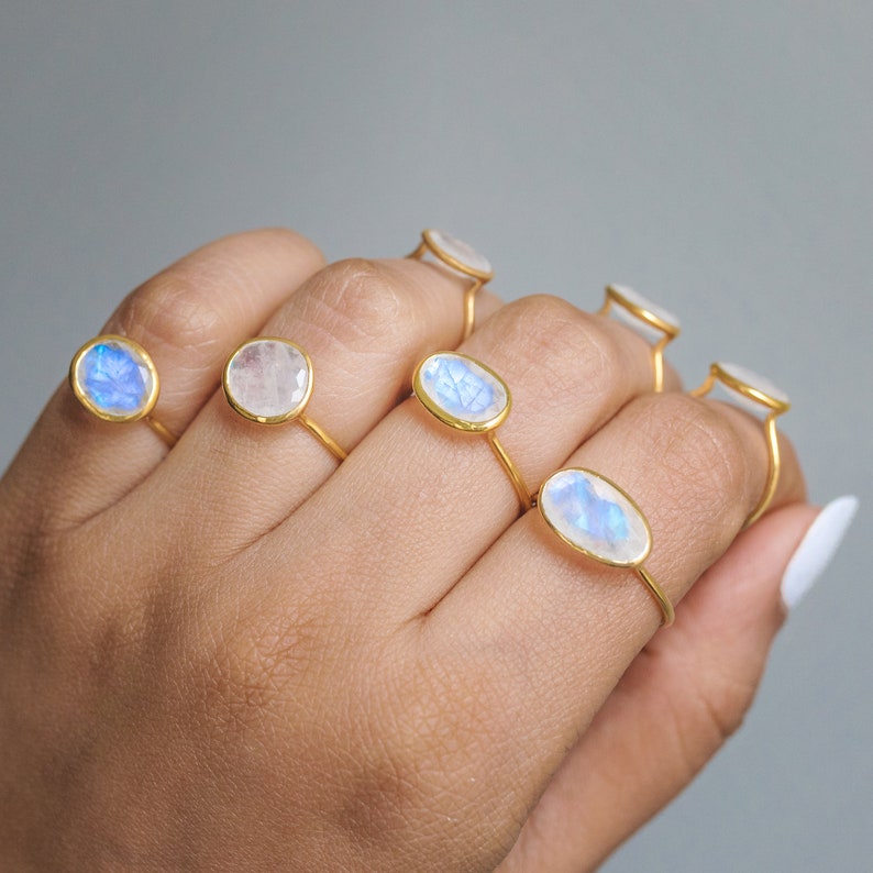 K&ouml;nnte beinhalten: Eine Sammlung goldener Ringe, jeder mit einem Mondstein in unterschiedlicher Form. Die Mondsteine zeigen eine Reihe von Farben, darunter Blau und Wei&szlig;. Die Ringe werden auf einer Hand vor neutralem Hintergrund pr&auml;sentiert.