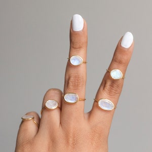 K&ouml;nnte beinhalten: Sechs goldfarbene Ringe mit ovalen, schillernden Mondsteinen. Die Ringe werden auf Fingern vor einem neutralen grauen Hintergrund pr&auml;sentiert. Die Ringe haben ein zartes, minimalistisches Design.