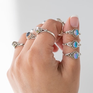 Pu&ograve; includere: Una mano che mostra diversi anelli d'argento con pietre preziose ovali iridescenti. Gli anelli hanno fasce ornate e dettagliate. Le pietre preziose mostrano una gamma di colori, tra cui blu, viola e verde. Lo sfondo &egrave; bianco.