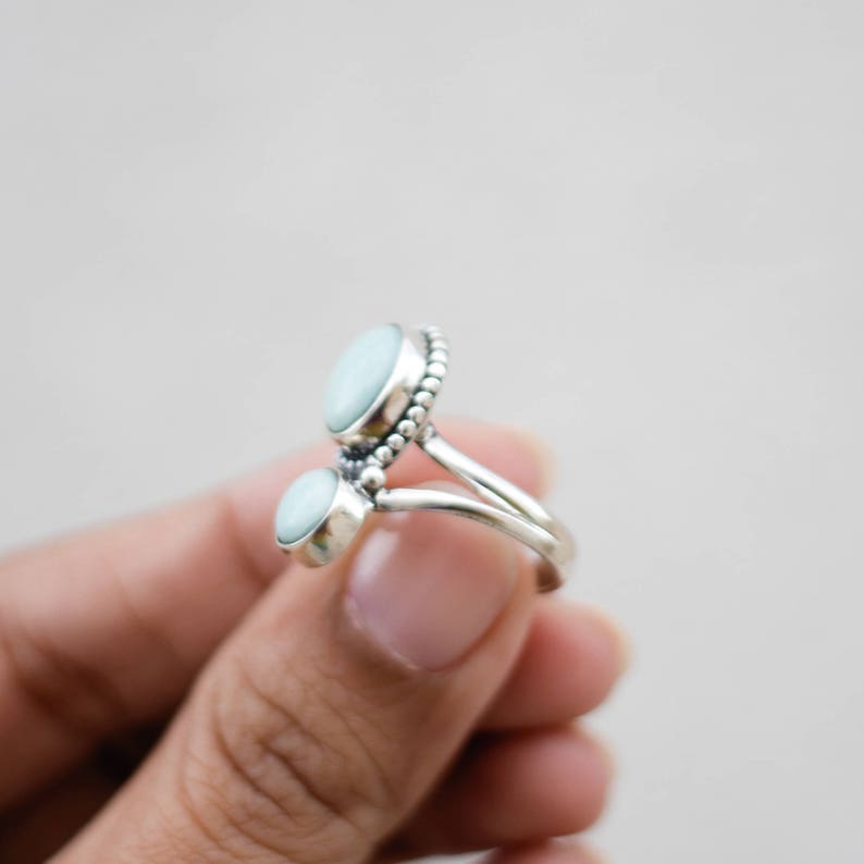 May include: A silver ring featuring two oval, light blue gemstones. The ring has a split shank design and a beaded border around the larger gemstone. The ring is held in a hand against a neutral background.