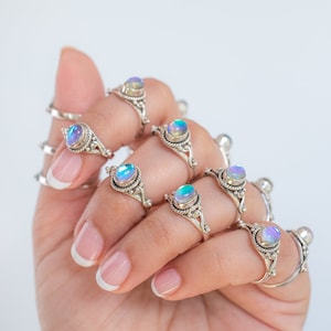 Pu&ograve; includere: Una mano mostra diversi anelli d'argento con pietre preziose ovali e iridescenti. Gli anelli hanno montature ornate in stile filigrana. Le pietre preziose cambiano colore, mostrando sfumature di blu, verde e viola, creando un effetto accattivante.