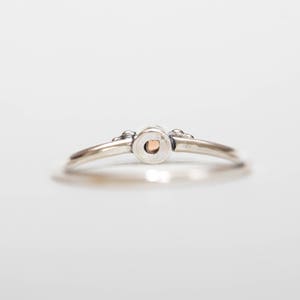 May include: A silver ring with a small, round, peach-colored stone set in the center. The ring has a delicate band and small decorative elements on either side of the stone setting. The ring is shown against a white background.