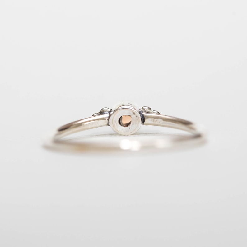 May include: A silver ring with a small, round, peach-colored stone set in the center. The ring has a delicate band and small decorative elements on either side of the stone setting. The ring is shown against a white background.