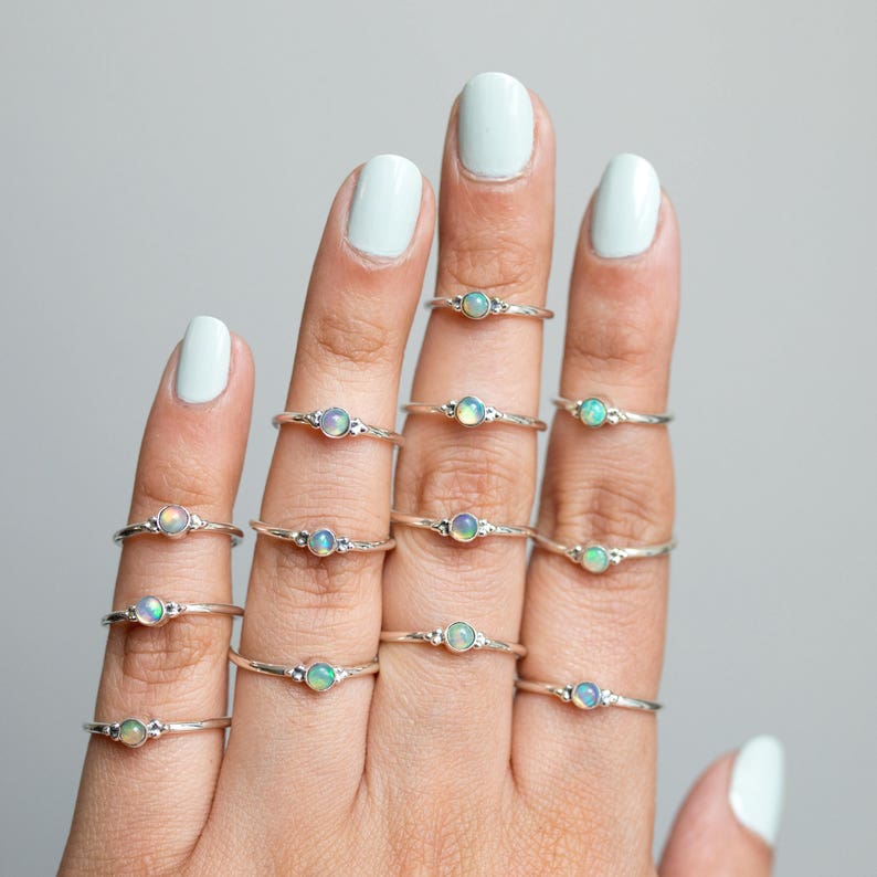 May include: A collection of delicate silver rings, each featuring a small, round opal gemstone. The rings are displayed on a hand with light blue nail polish. The opals display a range of colors, including white, blue, and green.