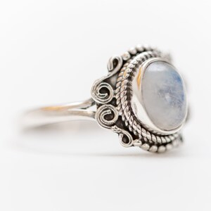 May include: A silver ring with an oval, light-colored gemstone. The ring has a detailed design with rope-like and swirl patterns around the gemstone. The band is smooth and polished.