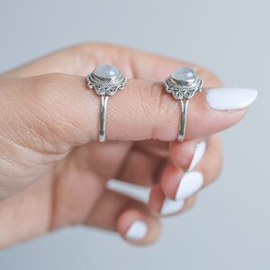 May include: Two silver rings with a round, light-colored stone set in an ornate bezel. The rings are displayed on a finger, showcasing the intricate details of the setting and band. The rings have a vintage or bohemian style.