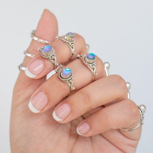 Pu&ograve; includere: Una mano mostra diversi anelli d'argento con pietre preziose ovali e iridescenti. Gli anelli hanno montature ornate e dettagliate. Le pietre preziose cambiano colore, mostrando sfumature di blu, viola e verde. Gli anelli sono in stile boh&eacute;mien.