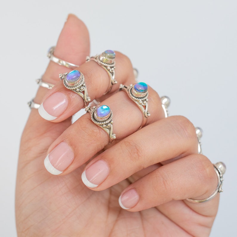 Pu&ograve; includere: Una mano mostra diversi anelli d'argento con pietre preziose ovali e iridescenti. Gli anelli hanno montature ornate e dettagliate. Le pietre preziose cambiano colore, mostrando sfumature di blu, viola e verde. Gli anelli sono in stile boh&eacute;mien.