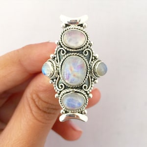 Peut inclure: Bague argent&eacute;e orn&eacute;e de cinq cabochons ovales en pierre de lune. La bague pr&eacute;sente un design orn&eacute; avec des volutes d&eacute;coratives et des d&eacute;tails en forme de corde autour des pierres. Les pierres de lune affichent un jeu de couleurs subtil.