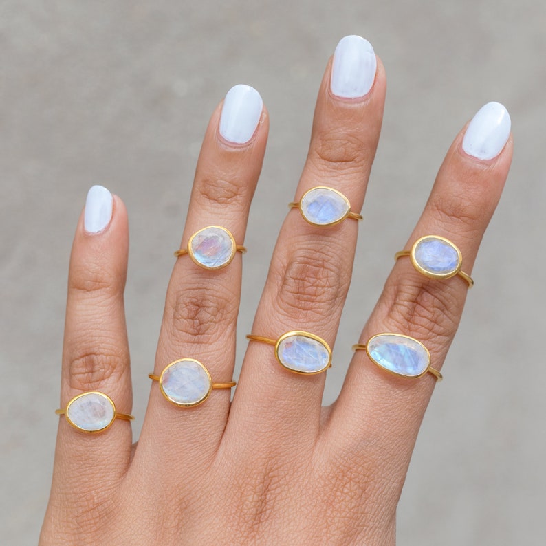 K&ouml;nnte beinhalten: Nahaufnahme einer Hand mit sieben goldfarbenen Ringen, jeder mit einem einzigartigen, ovalen oder runden, schillernden Edelstein. Die Edelsteine haben eine milchig-wei&szlig;e und blaue Farbe. Der Hintergrund ist hellgrau.