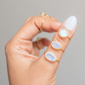 K&ouml;nnte beinhalten: Drei goldene Ringe mit ovalen, schillernden Mondstein-Cabochons werden auf einem Finger gezeigt. Die Ringe haben ein schlichtes, zartes Design. Ein vierter, d&uuml;nnerer Goldring ist ebenfalls sichtbar. Der Hintergrund ist einfarbig grau.