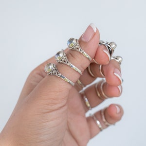 Pu&ograve; includere: Una mano mostra diversi anelli d'argento con pietre preziose rotonde e iridescenti. Gli anelli hanno fasce ornate in stile filigrana. Sono visibili anche anelli d'argento lisci aggiuntivi. Lo sfondo &egrave; bianco neutro.