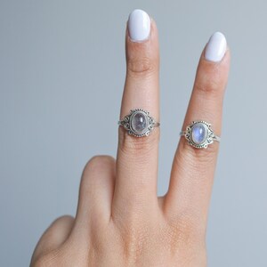 May include: Two silver rings with oval gemstones are displayed. The rings feature ornate, filigree-style settings with a beaded border. One ring has a gray gemstone, and the other has a blue-white gemstone. The rings are shown on fingers against a light gray background.