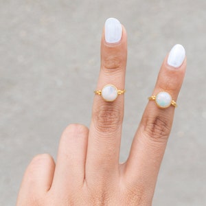 May include: Two gold rings, each featuring a round, iridescent opal stone. The rings are displayed on fingers with white nail polish against a neutral background. The rings have a delicate, minimalist design.