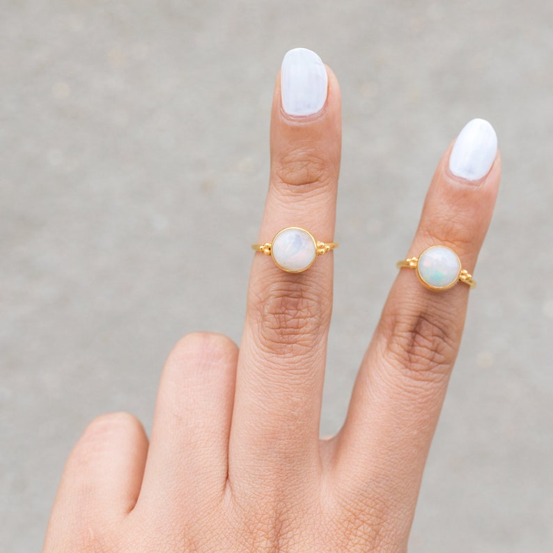 May include: Two gold rings, each featuring a round, iridescent opal stone. The rings are displayed on fingers with white nail polish against a neutral background. The rings have a delicate, minimalist design.