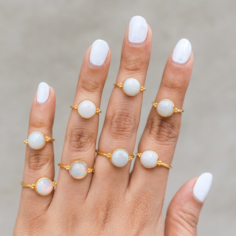 May include: A collection of gold-colored rings, each featuring a large, round, white opal stone. The rings are displayed on a hand with white nail polish. The rings have a simple, elegant design, perfect for everyday wear.
