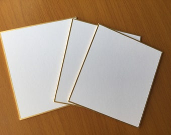 May include: A collection of white square cards with gold edging. The cards are displayed on a wooden surface, with three cards visible. The gold trim provides a touch of elegance. Suitable for invitations or stationery.