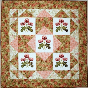 Könnte beinhalten: Eine Steppdecke mit einem Blumenmuster. Die Steppdecke besteht aus Quadraten mit weißem Hintergrund und einem Blumendesign in der Mitte. Die Quadrate sind von einem Rand aus Rosa und Grün umgeben.