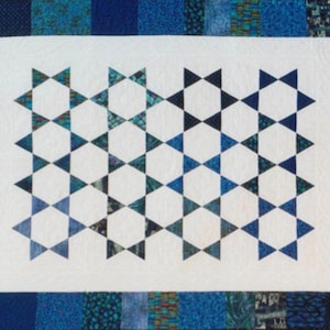 Puede incluir: Un edredón hecho a mano con un centro blanco que presenta un patrón de estrellas geométricas en tonos de azul y verde azulado. El edredón está bordeado con varias telas estampadas azules, creando un contraste visualmente atractivo.