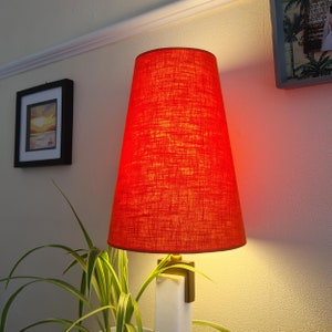 May include: A red lampshade with a textured fabric finish sits on a white marble base. The lampshade is lit and casts a warm glow.