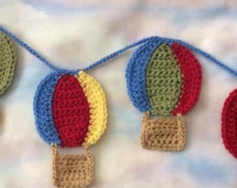 May include: A garland of crocheted hot air balloons in blue, red, yellow, and green. The balloons are strung together with a white cord.