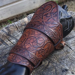 May include: A brown leather arm guard with intricate Celtic knotwork design, secured with straps. The arm guard is on a black hand, displayed on a weathered wooden surface. The leather has a rich, textured appearance.