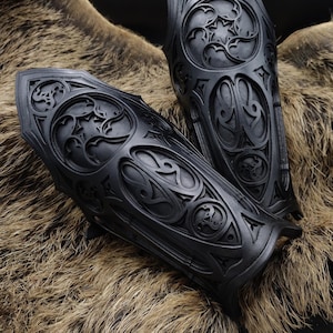 May include: A pair of black leather arm guards with intricate, carved designs. The guards are made of thick leather and have a medieval or fantasy aesthetic.