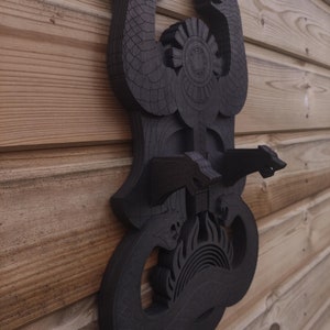 Pu&ograve; includere: Un'appendice da parete in legno nero con un design di due serpenti intrecciati attorno a un simbolo solare. I serpenti hanno squame dettagliate e il sole ha un design circolare con raggi che ne emanano.