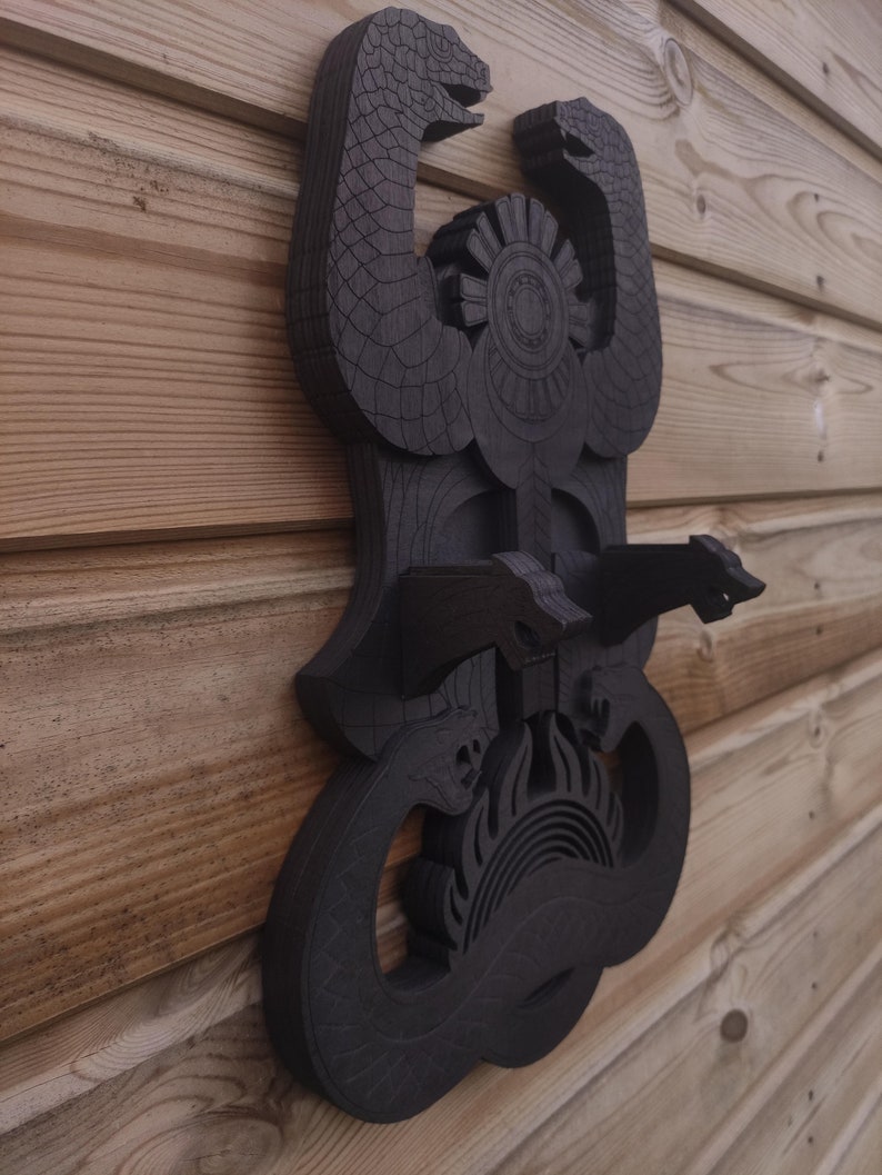 Pu&ograve; includere: Un'appendice da parete in legno nero con un design di due serpenti intrecciati attorno a un simbolo solare. I serpenti hanno squame dettagliate e il sole ha un design circolare con raggi che ne emanano.