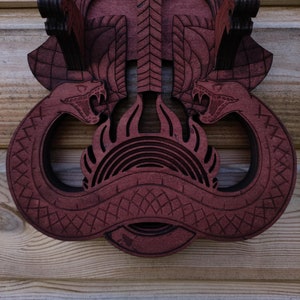 Pu&ograve; includere: Un'appendice murale in legno con un design intagliato di due serpenti con le teste rivolte l'una verso l'altra. I serpenti sono intrecciati e hanno un design a fiamma al centro. Il legno &egrave; di colore rosso scuro.
