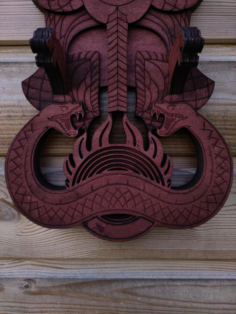 Pu&ograve; includere: Un'appendice murale in legno con un design intagliato di due serpenti con le teste rivolte l'una verso l'altra. I serpenti sono intrecciati e hanno un design a fiamma al centro. Il legno &egrave; di colore rosso scuro.