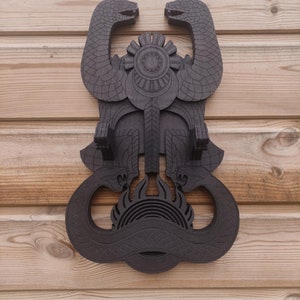 Pu&ograve; includere: Un supporto da parete in legno marrone scuro a forma di due serpenti intrecciati con un design circolare al centro. I serpenti hanno squame dettagliate e il design circolare ha un motivo a forma di sole.