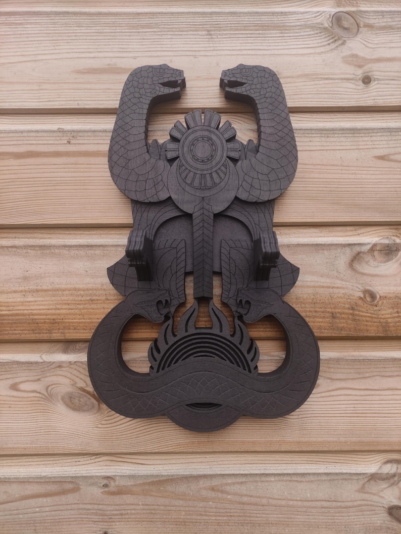 Pu&ograve; includere: Un supporto da parete in legno marrone scuro a forma di due serpenti intrecciati con un design circolare al centro. I serpenti hanno squame dettagliate e il design circolare ha un motivo a forma di sole.