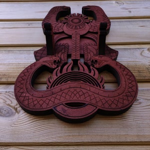 Pu&ograve; includere: Un'appendice murale in legno con un design rosso e nero di due serpenti intrecciati attorno a un simbolo solare. Il design &egrave; intricato e dettagliato, con un motivo geometrico sui corpi dei serpenti.