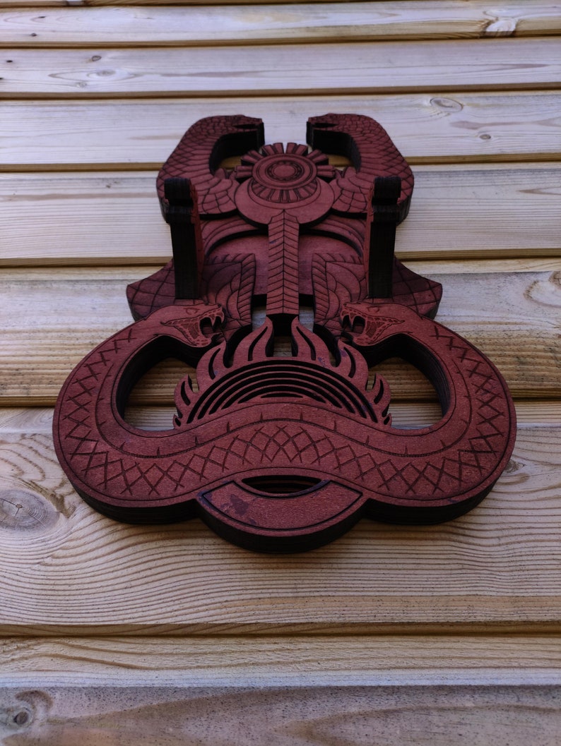 Pu&ograve; includere: Un'appendice murale in legno con un design rosso e nero di due serpenti intrecciati attorno a un simbolo solare. Il design &egrave; intricato e dettagliato, con un motivo geometrico sui corpi dei serpenti.