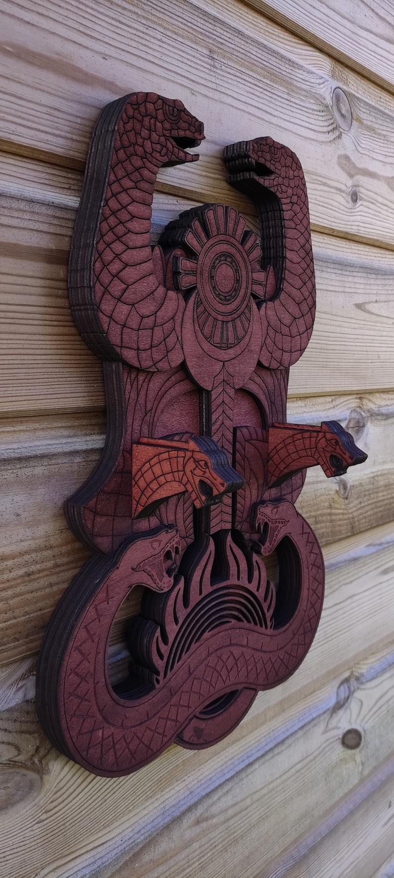 Pu&ograve; includere: Un'appendice murale in legno con un design a strati di serpenti intrecciati e un simbolo solare centrale. I serpenti sono scolpiti con squame e hanno la bocca aperta. Il simbolo solare &egrave; circondato da un design circolare.