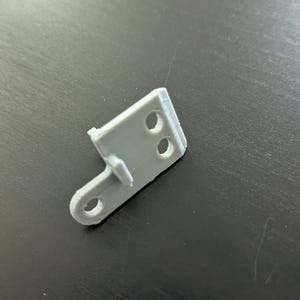 以下が含まれることがあります： 側面に2つの穴と小さなタブが付いた、グレーのプラスチック製の3Dプリントブラケットです