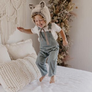 May include: A child wearing a brown and white faux fur bear hat jumps on a white bed with a cream-colored knitted throw blanket. The child is wearing a white t-shirt and blue denim overalls.